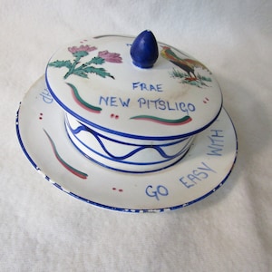 Pode incluir: Um pote de manteiga de cerâmica branca com um design azul e verde. A tampa apresenta um galo e o texto "FRAE NEW PITSLIGO". A base tem o texto "GO EASY WITH".