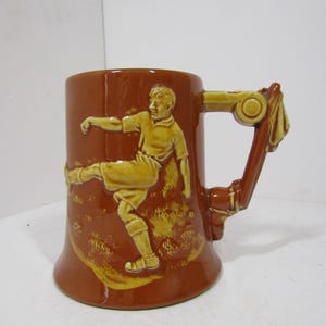 May include: A brown ceramic mug featuring a raised, light-colored relief of a soccer player in motion. The player is depicted mid-kick, wearing a jersey and shorts. The mug has a uniquely shaped handle that incorporates a decorative scroll design.