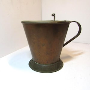 May include: A conical copper vessel with a dark, weathered patina, featuring a wide top rim, a narrow base, and a single curved handle attached to the side. A small metal loop is positioned on the top edge, providing a functional detail to the object.
