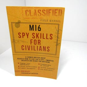 Peut inclure: Un livre de poche jaune intitulé M16 Spy Skills for Civilians, Field Manual. La couverture présente du texte noir et rouge, incluant un tampon classifié, une liste de techniques d'espionnage, l'auteur Red Riley et une préface d'Ian Sharp.