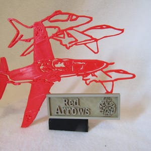 May include: Red Arrows, a 3D printed model of a formation of five red jet airplanes, with a plaque that reads "Red Arrows" and the Royal Air Force logo.