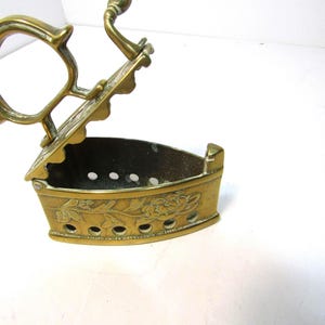 May include: A vintage brass sad iron with a hinged lid and a decorative floral relief pattern on the side. The metal body features a series of circular ventilation holes along the base and a curved handle attached to the top lid mechanism.