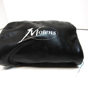 Puede incluir: Bolsa de cuero negro con el texto blanco "Motens" en una fuente estilizada. La bolsa es rectangular con bordes redondeados y parece estar hecha de un material suave y flexible. El fondo es blanco.