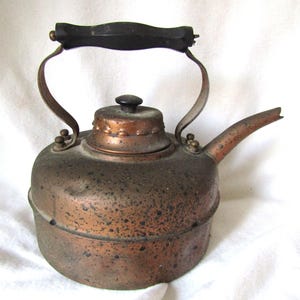 Può includere: Un bollitore di rame vintage con un manico in legno nero e una finitura in rame ossidato. Il bollitore ha un corpo arrotondato e un beccuccio laterale.