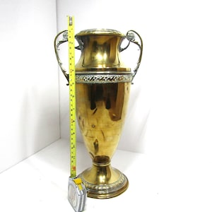 May include: A large, antique brass urn with ornate handles and decorative trim. A yellow measuring tape shows the urn is approximately 24 inches tall. The urn has a wide base and a flared top, with a polished finish.