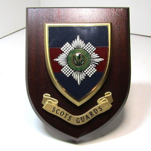 Pode incluir: Placa de madeira com um emblema em forma de escudo. O emblema apresenta uma borda dourada, um fundo azul e vermelho e as palavras "SCOTS GUARDS" em uma faixa dourada.