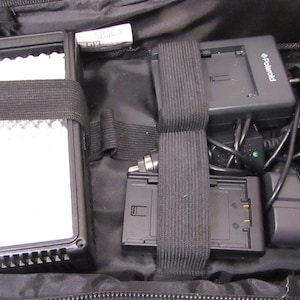 Puede incluir: Bolsa de cámara negra que contiene un panel de luz, un cargador Polaroid y cargadores de batería. La bolsa tiene correas negras para asegurar los artículos. Un paquete de sílice es visible dentro de la bolsa.