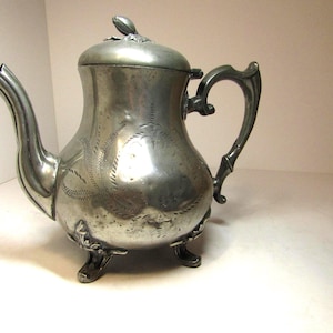 Op de afbeelding: Een zilveren theepot met een versierd ontwerp en drie poten. De theepot heeft een deksel met een decoratieve knop.