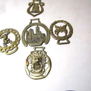 May include: A collection of antique brass horse brasses. The designs include a bell, a church, a butterfly, a lion, and a horse. These decorative pieces are likely from the 19th or early 20th century.