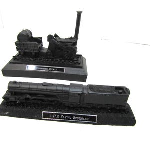 Puede incluir: Dos modelos de trenes en miniatura de color negro sobre bases rectangulares. El modelo superior es el Stephenson's Rocket con una chimenea alta y un vagón pequeño. El modelo inferior es la locomotora de vapor 4472 Flying Scotsman con ténder, cada uno con una placa.