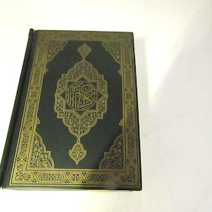 May include: A dark blue hardcover book featuring intricate gold foil stamping on the front cover. The design includes a central medallion with Arabic calligraphy surrounded by ornate geometric and floral patterns along the rectangular border.