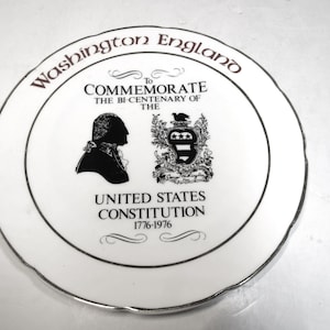 May include: A white commemorative plate with a silver rim. The plate features text that reads "Washington England" and "To Commemorate The Bi-Centenary of The United States Constitution 1776-1976". The plate has a black silhouette of a man and a coat of arms.