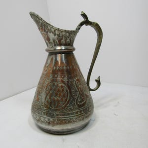 May include: An antique copper and silver-toned jug with an ornate design. The jug features a decorative handle and spout, with etched patterns covering the body. The metal jug is likely used for serving beverages.