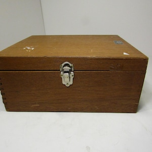 Puede incluir: Una caja rectangular de madera con tapa con bisagras y cierre metálico. La caja es de madera marrón claro y tiene un diseño sencillo y clásico. La caja está cerrada y parece vacía.