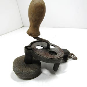 Könnte beinhalten: Ein antiker handgekurbelter Schleifstein mit Holzgriff. Die Metallkomponenten weisen Gebrauchsspuren auf. Der Stein hat eine abgerundete, graue Farbe. Der Kurbelmechanismus ist an einer kreisförmigen Metallplatte befestigt.