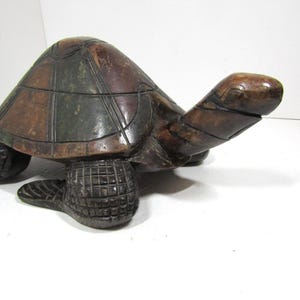 Könnte beinhalten: Eine dunkelbraune und grüne geschnitzte Holzschildkrötenfigur mit segmentiertem Panzermuster und strukturierten Beinen. Die Schildkröte liegt neben einem gelben Maßband, das eine Länge von etwa 30 cm von Kopf bis Schwanz anzeigt.