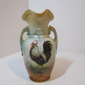 Peut inclure: Un vase en porcelaine avec un bord festonné et deux anses, orné d'un coq peint à la main avec une crête rouge, un cou blanc et des plumes de queue noires, debout dans un champ herbeux sous un ciel bleu clair, avec des finitions dorées.