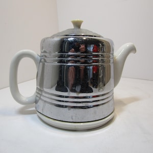May include: A vintage chrome teapot with a white handle, spout, and base. The teapot has a reflective surface and a clear lid with a small, light-yellow knob. The design features horizontal bands around the body.