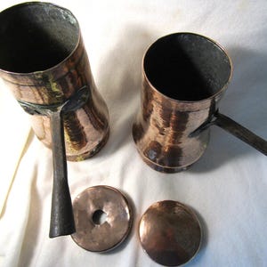 May include: Two cylindrical copper vessels with dark metal handles extending from the sides. Each vessel has a matching circular copper lid, one of which features a central hole. The copper surfaces exhibit a hammered texture and warm tones.