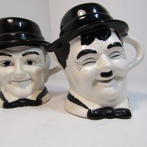 May include: Two ceramic mugs shaped like the faces of Laurel and Hardy. Each mug is white with black accents, including a bowler hat, bow tie, and facial features. The mugs have handles.