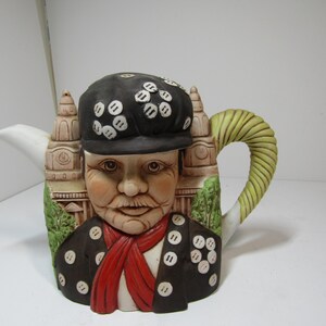 Może przedstawiać: Ceramiczny czajniczek w kształcie postaci w czarnej czapce i kurtce ozdobionej białymi guzikami, czerwonym szalikiem i zielonym kołnierzem. Tło przedstawia beżową konstrukcję budynku, a uchwyt ma formę teksturowanej, jasnozielonej spirali.