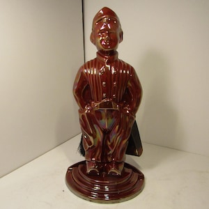 Puede incluir: Figura de cerámica vintage de un hombre con uniforme militar, de pie firme. La figura es de un marrón rojizo intenso con reflejos iridiscentes. Tiene una base redonda y una borla negra.