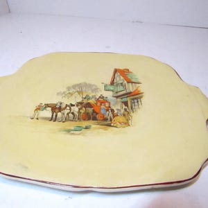 May include: A cream-colored ceramic serving platter with a scalloped edge and a brown rim. The center features a colorful illustration of a horse-drawn stagecoach arriving at a building with a red roof, surrounded by several figures in period clothing.