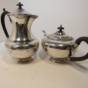 May include: A silver tea set with a teapot and a creamer. The teapot has a curved spout and a black handle. The creamer has a tall, curved body and a black handle. Both pieces have decorative detailing.