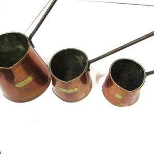 Puede incluir: Tres ollas antiguas de cobre con largos mangos de metal oscuro. Cada olla tiene un cuerpo redondeado y una pequeña etiqueta rectangular de latón. Las ollas están dispuestas de mayor a menor, mostrando sus diferentes tamaños.