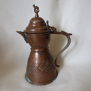 May include: A copper teapot with an ornate handle and a lid featuring a small camel finial. The teapot has an engraved design and a patina finish.