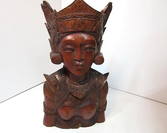 May include: A carved wooden bust of a woman with an ornate headdress and detailed carvings. The bust is a reddish-brown colour and features a serene facial expression. The figure has decorative shoulder pieces and a detailed bodice.