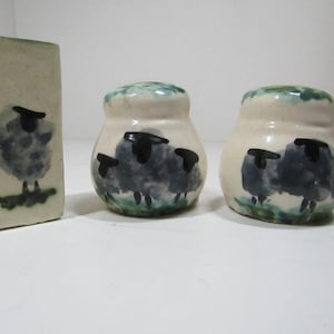 Può includere: Un set di tre pezzi in ceramica composto da un blocco rettangolare e due spargitori arrotondati, tutti caratterizzati da illustrazioni dipinte a mano di pecore grigie soffici con teste nere su fondo color crema con accenti verdi alla base.