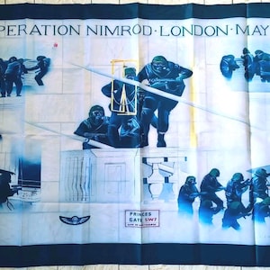 Op de afbeelding: Een rechthoekige stoffen banner met een blauw-witte illustratie die een militaire operatie uitbeeldt. De tekst "OPERATION NIMROD - LONDON - MAY 5th 1980" staat bovenaan. De onderkant heeft de tekst "PRINCES GATE SW7".