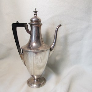 May include: A silver teapot with a black handle and a monogrammed design. The teapot has a round body and a tall spout.