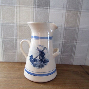 Puede incluir: Una jarra de cerámica blanca con un diseño de molino de viento azul y blanco. La jarra tiene dos asas y un pico.