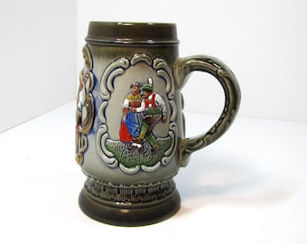 May include: A ceramic beer stein featuring a dark olive green rim and base with a central relief panel depicting a couple in traditional attire dancing on a green field. The stein has a curved handle and a light grey body with ornate borders.