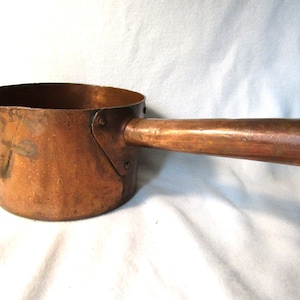 Könnte beinhalten: Ein antiker Kupfertopf mit einem langen Holzgriff. Der Topf hat einen abgerundeten Boden und einen ausgestellten Rand. Der Griff ist mit zwei Nieten am Topf befestigt.