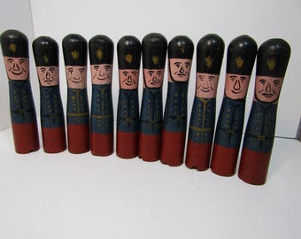 Op de afbeelding: Een verzameling van negen houten speelgoedsoldaten. Elke soldaat is met de hand beschilderd met een zwarte hoed, een blauwe jas en een rode basis. De soldaten hebben geschilderde gezichten met glimlach.