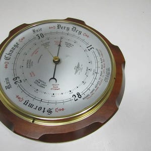 Könnte beinhalten: Ein rundes Wandbarometer mit einem dunkelbraunen Holzrahmen und einer messingfarbenen Lünette. Das silberne Zifferblatt zeigt schwarzen Text wie Stormy, Rain, Change, Fair und Very Dry sowie numerische Skalen und zwei schwarze Metallzeiger.