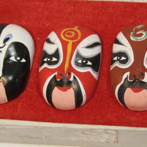 May include: Three decorative, oval-shaped masks with painted faces. The masks are in black, white, red, and brown, with detailed facial features. They are displayed on a red surface.