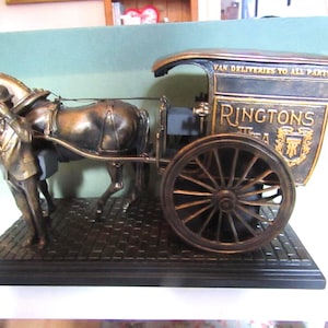 May include: A bronze-toned figurine depicting a horse-drawn delivery cart with a driver standing alongside. The cart features the text "RINGTONS TEA" and "VAN DELIVERIES TO ALL PARTS" with a crest emblem, all mounted on a rectangular brick-patterned base.