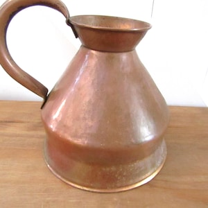 Könnte beinhalten: Eine antike Kupferkanne mit breiter Basis und einem ausgestellten Rand. Die Kanne hat einen einzigen Henkel und eine abgenutzte, gealterte Patina.