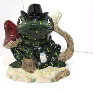 Puede incluir: Una caprichosa figura de rana de cerámica verde con lunares amarillos, con un sombrero negro. Tiene una seta roja y blanca y una bolsa marrón. La base dice "SURGERY". La rana sostiene una pequeña botella.