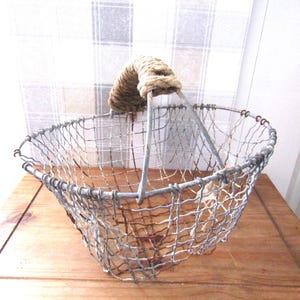 Puede incluir: Una cesta de alambre vintage con un asa de cuerda. La cesta está hecha de alambre metálico y tiene un aspecto rústico y desgastado.