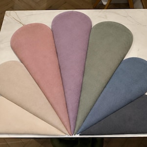 Op de afbeelding: Een waaier van zeven druppelvormige kussens in beige, roze, lavendel, olijfgroen, blauw en antracietgrijs. De kussens zijn gerangschikt op een wit oppervlak met marmerlook.