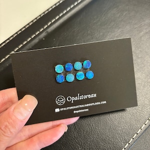 May include: A black card with eight round blue opal cabochons arranged in two rows of four. The card has the text "Opalstoreau" and the email address "opalstoreaustralia@outlook.com" and the social media handle "@opalstoreau".