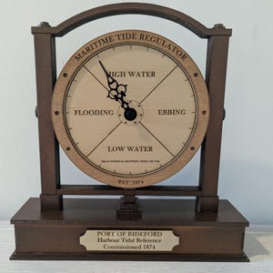 Op de afbeelding: Een houten Maritime Tide Regulator met een ronde wijzerplaat die de getijdenstanden aangeeft: Hoog Water, Vloed, Eb en Laag Water. De klok heeft een donkerbruin frame en voet, met de tekst "Port of Bideford, Harbour Tidal Reference, Commissioned 1874."