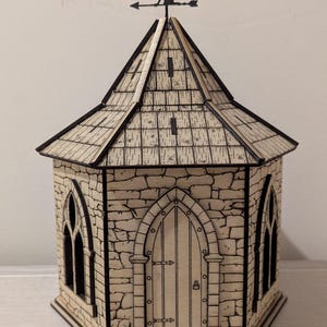 Può includere: Una scatola ottagonale in legno con facciata a forma di pietra. La scatola ha un tetto a capanna e finestre e porte ad arco. Un'altana con la sagoma di una fata è sul tetto. La scatola è in legno chiaro con linee incise scure.