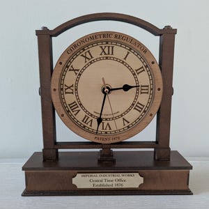 May include: Wooden chronometric regulator clock with Roman numerals and black hands. The clock face is light brown, and the frame is dark brown. The base has a plaque that reads "IMPERIAL INDUSTRIAL WORKS Central Time Office Established 1876."