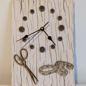 May include: A white wooden clock with a black clock face and hands. The clock is decorated with brown buttons and a gold measuring tape and scissors.